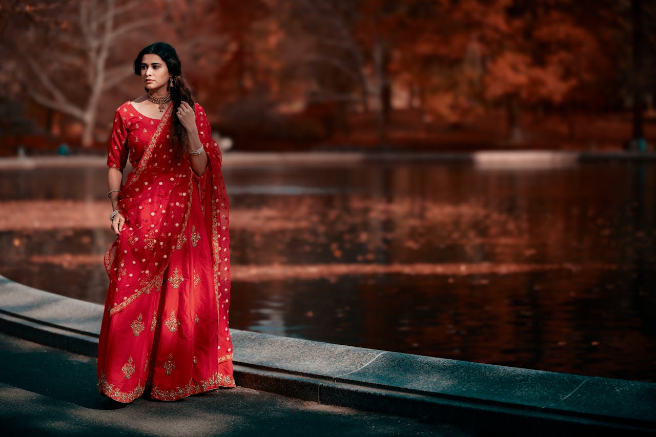An elegant woman wearing a traditional red saree walks by a picturesque lake in the park during the autumn season.