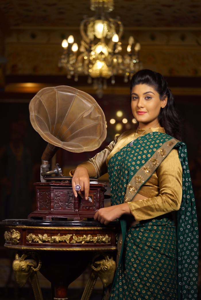 Smiling young Indian female in Sari dress leaning on old fashioned gramophone while standing in room with crystal chandelier